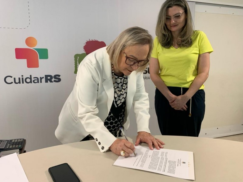 Foto em plano americano da secretária Arita inclinada sobre uma mesa branca assinando um documento. Atrás dela, uma mulher está de pé observando o ato.