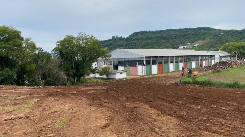 Imagem de uma área já limpa após retirada dos resíduos da enchente. Trata-se de um terreno limpo com uma construção que parece ser de uma escola ao fundo.