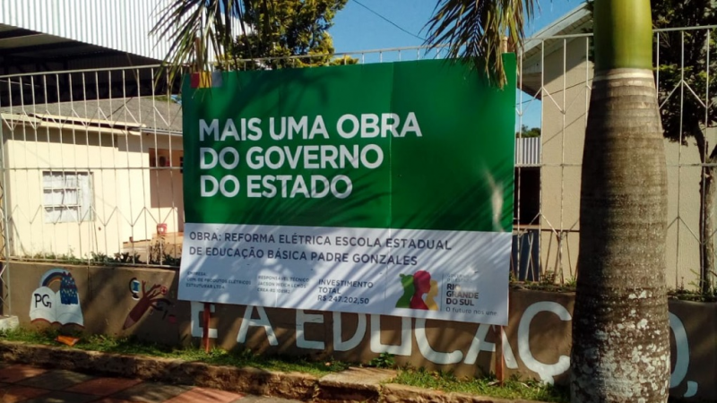Foto em ambiente aberto da fachada da escola, onde está uma grande branca com uma grande placa verde na qual se lê "Mais uma obra do governo do Estado".