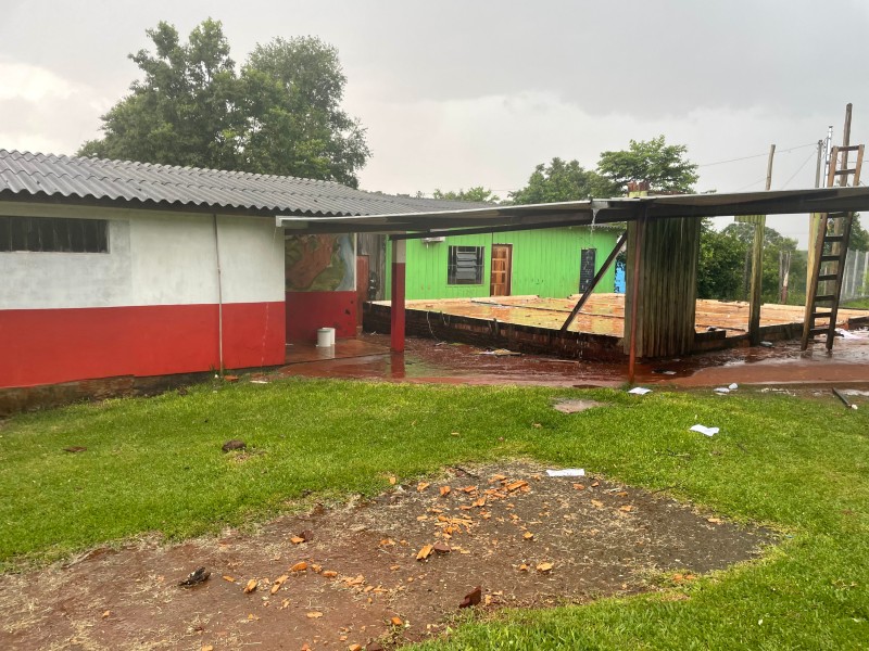 Foto em ambiente aberta do pátio da escola. Há uma parte de gramado e outra cimentada. A escola é pintada de branco e vermelho e possui lajes.