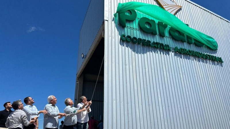 Foto em ambiente aberto. Seis homens puxam um cordão para desvelar a logo "Paraíso: Cooperativa Agroindustrial", de cor verde, no que parece ser um grande contêiner. Eles estão no canto inferior da esquerdo da foto, e a logo está no canto direito superior.