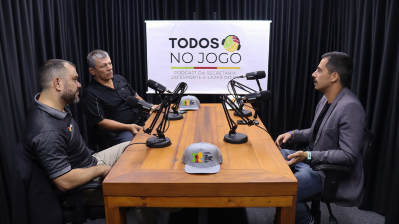 Foto em ambiente interno, em um estúdio de gravação. Ao redor da mesa, com um microfone para cada, estão três homens gravando o programa. Ao fundo, está a marca do podcast Todos no Jogo. A marca destaca o próprio nome e um desenho de uma bola, nas cores da bandeira do RS (verde, vermelho e amarelo), com um fone de ouvido ao redor. Abaixo, está escrito "Podcast da Secretaria do Esporte e Lazer do RS".