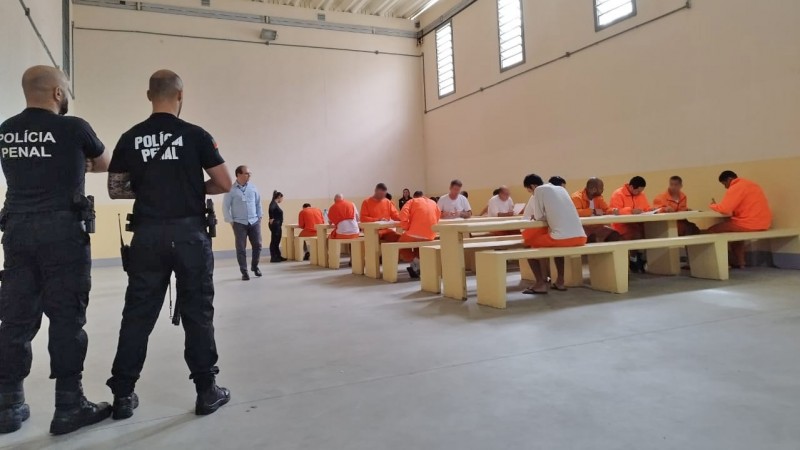 Imagem em ambiente interno, em uma penitenciária. Em primeiro plano, de costas, estão dois agentes penitenciários. Nas roupas que usam, lê-se "Polícia Penal". Mais ao fundo da foto, há diversas pessoas privadas de liberdade realizando as provas. Elas estão sentadas em bancos e mesas coletivas, vestidas com uniformes cor de laranja ou camisetas brancas. Eles olham para a prova, de cabeça baixa, concentrados.