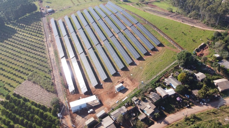 Imagem aérea de uma usina fotovotaica. Num ambiente rural, vários painéis inclinados, de cor escura, se encontram refletindo o sol.