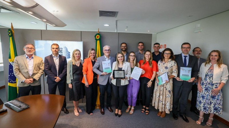 Foto em ambiente interno, em uma sala de reuniões. Quinze autoridades, homens e mulheres, estão alinhados em pé, lado a lado, posando para a câmera e mostrando o documento entregue. Ao fundo, há uma bandeira do Brasil e uma do RS.