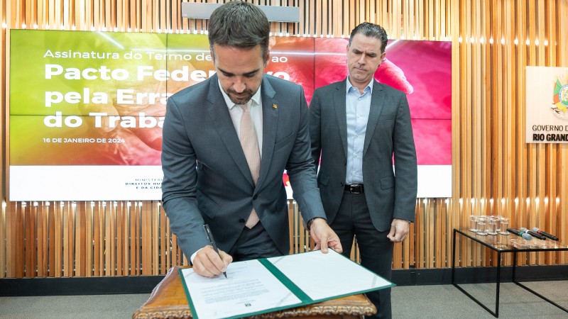 Foto em ambiente fechado. Leite aparece, em plano americano, assinando o documento. Ele está de pé, levemente inclinado sobre uma mesa com o papel. Ao fundo, Peruchin observa o ato.