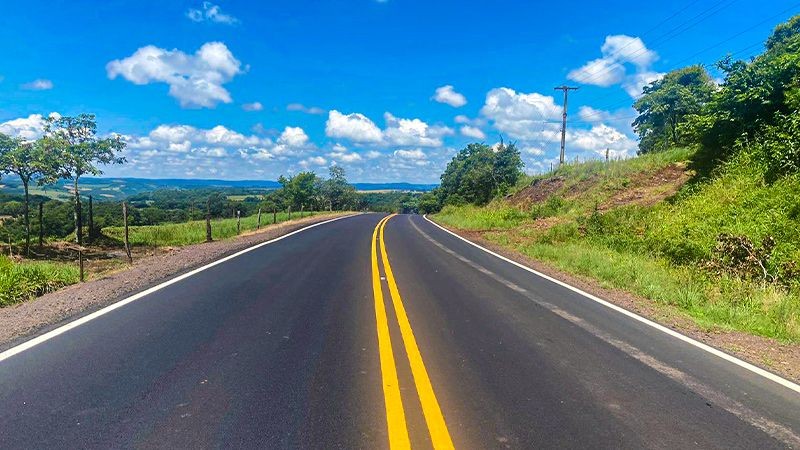Foto em ambiente aberta da estrada de mão dupla, em bom estado de conservação. O céu está azul com nuvens, e nos arredores há mato e árvores.