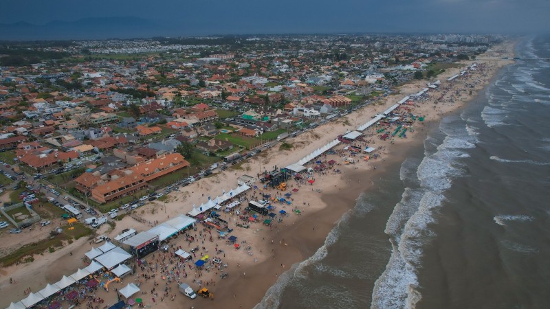 Foto aérea em ambiente aberto da beira da praia. À esquerda, está a parte das casas e da faixa de areia; à direita, o mar.