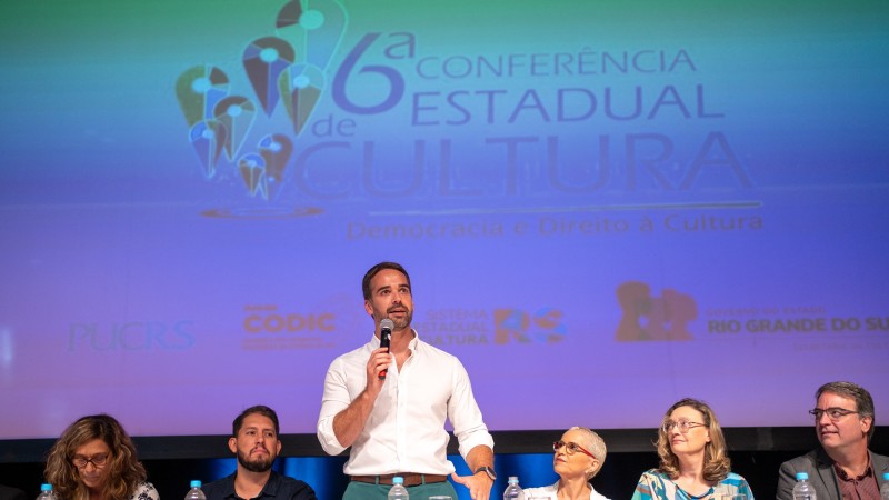 Leite em pé com o microfone e falando à plateia. Ele compõe a mesa do evento. Sentados à esquerda, uma mulher e um homem, e sentados à direita, duas mulheres e um homem. Atrás do governador há uma enorme tela onde se lê Sexta Conferência Estadual de Cultura.