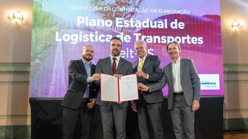 Foto posada em que Leite, ao centro e em pé, segura o documento assinado acompanhado pelo presidente da Infra S.A. e dos secretários de Parcerias e Concessões e de Logística e Transportes. Atrás deles há uma tela grande onde se lê: Plano Estadual de Logística de Transportes.