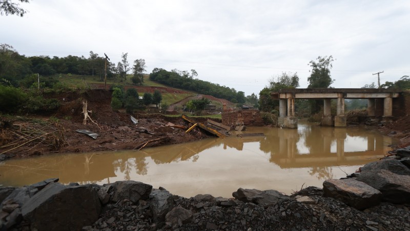 Imagem da ponte destruída e pela metade. Há água barrenta no rio e alguns destroços na parte que foi danificada.