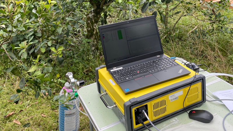 Foto em ambiente aberto. Numa região de mata, um notebook está sobre uma mesa e um aparelho amarelo retangular. Ao lado, há o que se assemelha a um extintor.