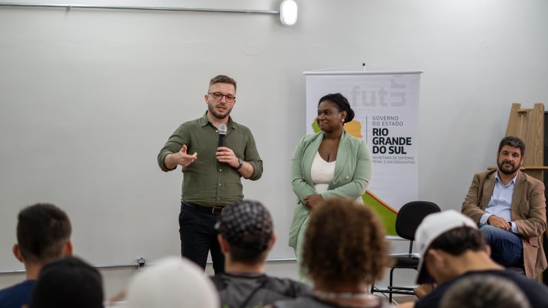 Foto em ambiente fechado de Cesar e Lisiane falando aos socioeducandos, que estão sentados (só é possível vê-los de costas, no primeiro plano da imagem). Ambos estão em pé. Cesar fala ao microfone.