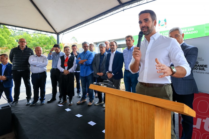 Foto em que Leite aparece meio de lado, à direita da imagem, discursando de pé junto ao púlpito. Com uma mão, ele segura o microfone; com a outra, gesticula. Ao fundo, vários homens enfileirados estão de pé acompanhando a fala.
