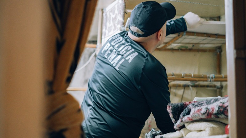 Foto em ambiente fechado de um agente, trajando roupas pretas com os escritos "Polícia Penal" nas costas. Ele está de costas para a câmera e vasculha pertences em uma cela.