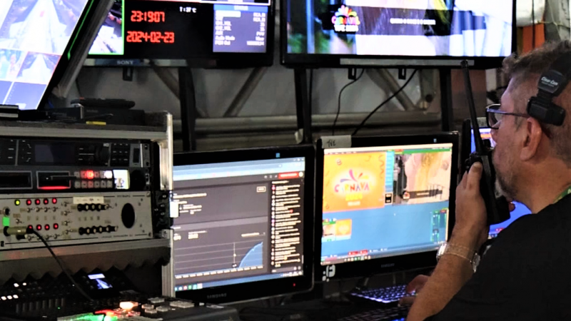 Um operador de vídeo de perfil, mais à direita da imagem, falando ao rádio e monitorando diversas telas e equipamentos na frente dele.