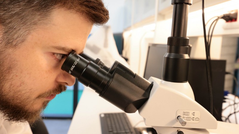 Foto de homem (que aparece à esquerda, enquadrado apenas na cabeça, de lado), observando algo através de um microscópio.