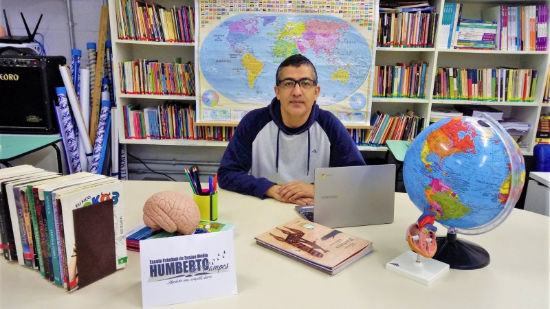 Foto, em sala fechada, do professor sentado junto a uma mesa, posando para a câmera. A mesa está com livros, notebook e um globo. Ao fundo, há uma grande estante de livros e um mapa mundi.
