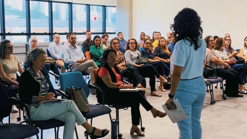 Num auditório, uma mulher aparece de pé, de costas para a câmera e de frente para a plateia, sentada em frente a ela em cadeiras. Cerca de 30 pessoas acompanham a fala.