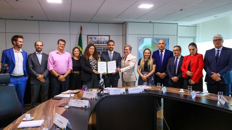 Foto posada. Leite e a ministra seguram o documento recém assinado, rodeado por outras pessoas que estava na reunião. Todos estão em pé atrás da mesa de reunião.