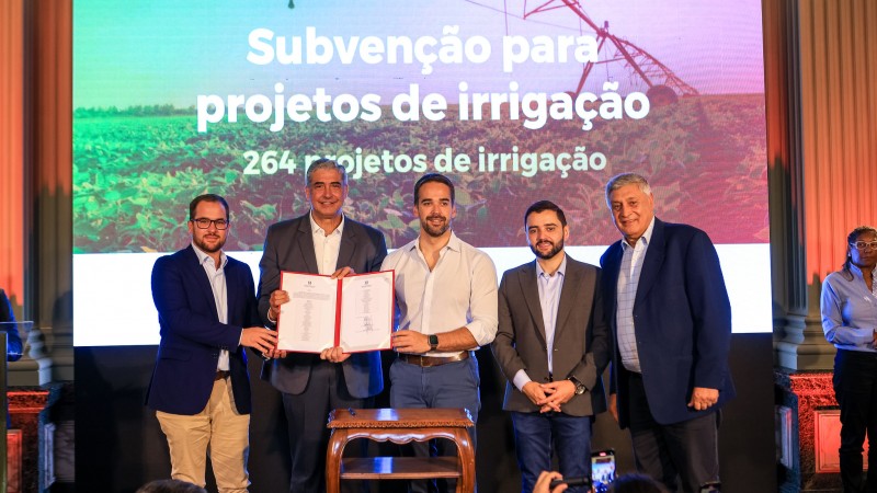 Foto posada, no palco montado no interior do Palácio Piratini, de Leite e mais quatro representantes do Estado sorrindo para a câmera enquanto seguram e mostram o documento assinado. Ao fundo, há um telão no qual está escrito "Subvenção para projetos de irrigação".
