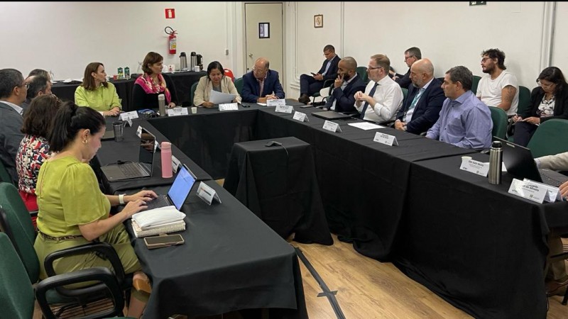 Imagem de uma mesa de reunião organizada em forma de U com diversas pessoas sentadas participando do encontro. Do lado direito, há uma fileira de cadeiras com algumas pessoas acompanhando a reunião.