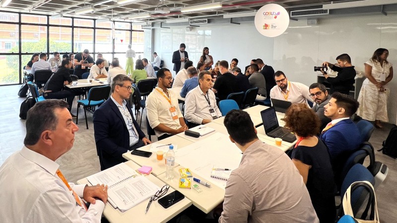 Imagem de uma grande sala com diversas mesas e pessoas sentadas conversando em grupos.