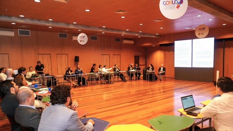 Imagem de uma grande sala com mesas organizadas em formato de u nas quais estão diversas pessoas sentadas debatendo. Mais à direita pode ser vista uma tela com uma projeção que está ilegível. Presos ao teto, podem ser vistos três displays redondos com a marca do Cosud.