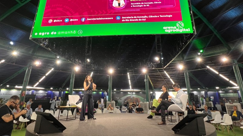 Imagem de uma grande palco no qual a secretária está em pé falando ao microfone e outras duas pessoas sentadas mais à direita. A plateia está em volta da arena e no alto há uma enorme tela em que é projetado o nome e o cargo da secretária Simone Stülp.