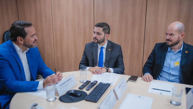 Sentados, conversando à mesa de reuniões, Gabriel Souza no centro, Capeluppi à direita e o secretário no Ministério dos Transportes à esquerda.