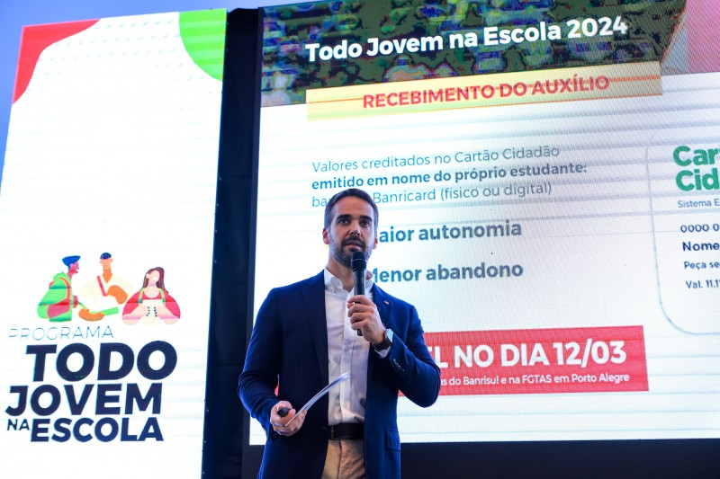 Leite em pé, da cintura para cima, falando ao microfone. Atrás dele há uma tela onde são projetadas informações sobre o programa. Mais à esquerda e em letras maiores está projetado: Todo Jovem na Escola.