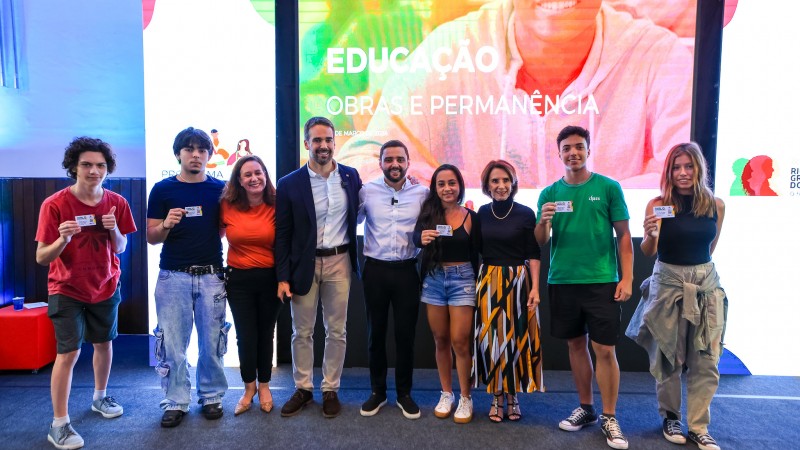 Foto posada. Em pé em um tablado estão Leite, Gabriel, Raquel Teixeira, cinco alunos que receberam o Cartão Cidadão do programa e a diretora da escola à qual eles pertencem.