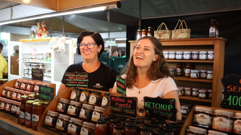 Duas mulheres que lideram empreendimento da agricultura familiar sorrindo atr&aacute;s de um balc&atilde;o cheio de produtos em um estande do Pavilh&atilde;o da Agricultura Familiar na Expodireto Cotrijal 2024