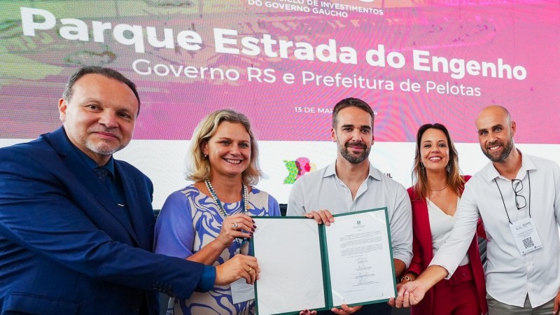 Leite e a prefeita de Pelotas seguram o documento que foi assinado em foto posada. Ao lado deles estão mais um homem do lado esquerdo e uma mulher e um home do lado direito. Ao fundo pode ser lido em um painel: Parque Estrada do Engenho. Governo do RS e Prefeitura de Pelotas.