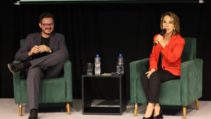 Raquel ao microfone mais à direita falando para plateia e Wislei Pereira mais à esquerda. Os dois estão sentados e participaram do encerramento da primeira Semana Estadual da Educação.