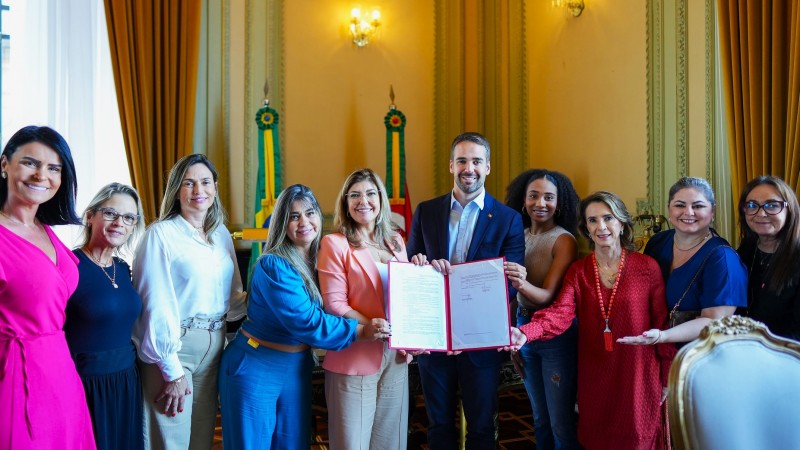 Foto posada. Leite ao centro junto com a deputada segurando uma pasta aberta com o documento da lei sancionada. Ao lado deles, três mulheres do lado esquerdo e quatro do lado direito.