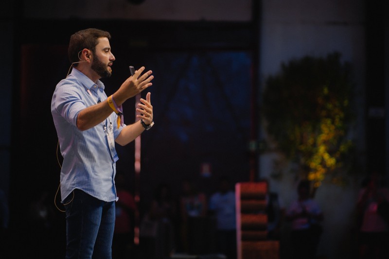 Em um palco, em ambiente fechado, Gabriel Souza gesticula com as mãos e fala para o público. A luz está direcionada para ele, enquanto o restante do ambiente está escuro.