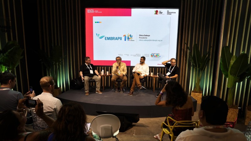 Quatro homens sentados sobre um pequeno tablado participando do painel. Atrás deles há uma tela na qual está projetada a palavra Embrapii. De costas a Plateia acompanha a fala dos painleistas.