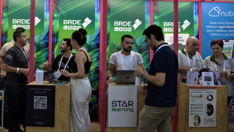 Espaço BRDE Labs II no South Summit Brazil 2024. Na imagem, há três pequenas bancadas com pessoas em pé dando explicações e outras acompanhando.