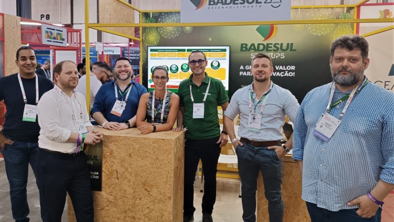 Foto posada na qual seis homens e uma mulher estão em pé diante do estande do Badesul no South Summit Baazil 2024.