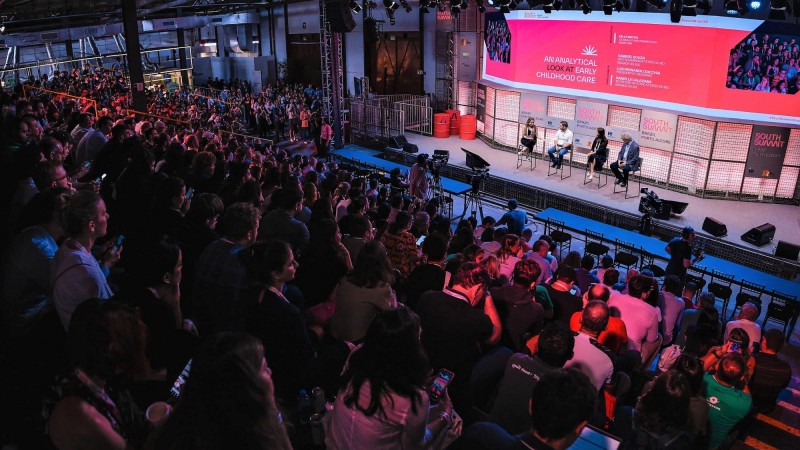 Imagem do alto do palco principal do South Summit Brazil mostrando boa parte da plateia de perfil mais à esquerda e ao centro e no canto superior direito os painelista sentados em cadeiras sobre um palco, com uma enorme tela atrás e sobre eles.