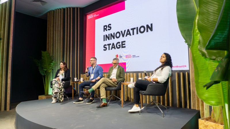 Duas mulheres e dois homens sentados em cadeiras sobre um tablado. Um deles fala ao microfone. Atr&aacute;s deles uma tela exibe as palavras RS Innovation Stage.