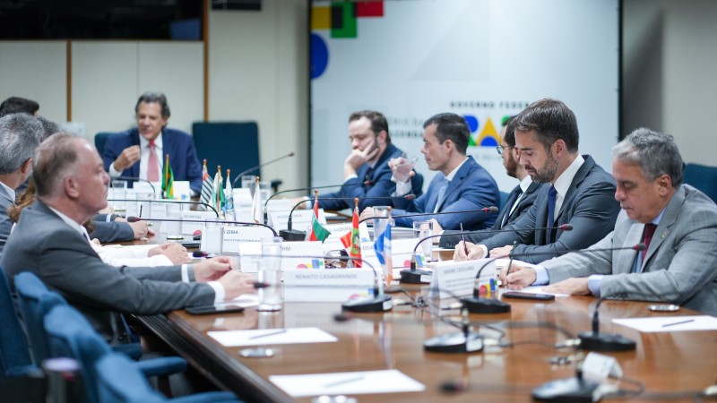 Numa sala de reunião, em torno de dez pessoas estão sentadas junto a uma grande mesa retangular. Na cabeceira está Haddad. Leite aparece à direita no quadro.