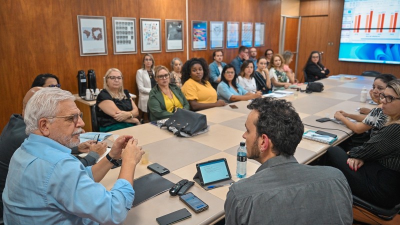 Imagem de uma reunião com diversas pessoas sentadas em torno de uma mesa grande e algumas outras sentadas em uma fileira de cadeiras encostada na parede. Há quadros em uma das paredes e uma tela na qual é projetado um gráfico em outra parede.