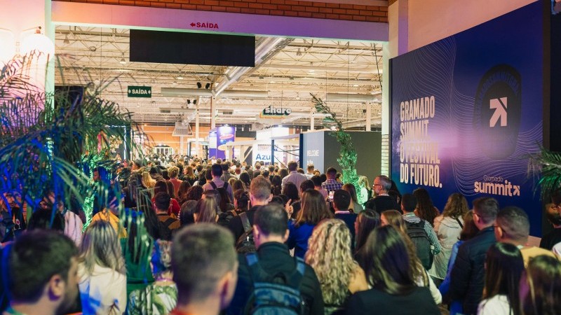 Imagem em ambiente interno, nos corredores do Gramado Summit. Ao lado direito, há um grande painel em cor roxa, com a seguinte inscrição em branco: "Gramado Summit - o festival do futuro". Ocupando todo o corredor, há uma multidão de pessoas, de costas, caminhando pelo evento.