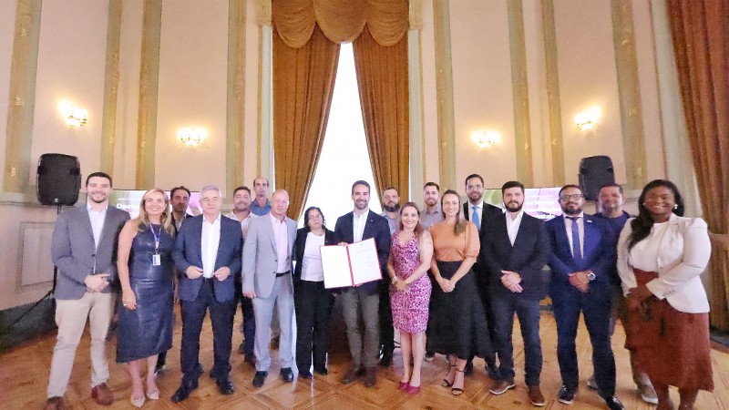 Foto em ambiente fechado. Ao centro da imagem, estão diversas autoridades em pé, posando para a câmera. Localizado em meio a elas, está o governador Eduardo Leite, segurando o documento assinado e sorrindo. Ao fundo, aparecem enormes janelas e cortinas douradas do Palácio Piratini.