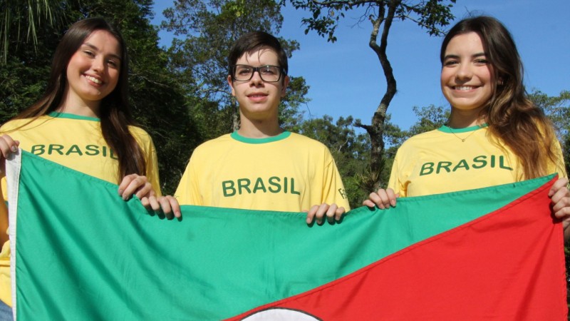 Os três estudantes estão em pé, ao ar livre, com árvores ao fundo, segurando a bandeira do RS e sorrindo para a câmera.