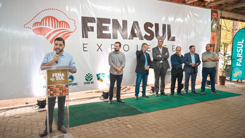 Foto de Gabriel Souza falando ao microfone, em pé, atrás de um púlpito, no lançamento da Fenasul Expoleite 2024. hÁ um enorme painel atrás dele com o nome da feira e seis homens sobre um tapete verde mais à direita da imagem.