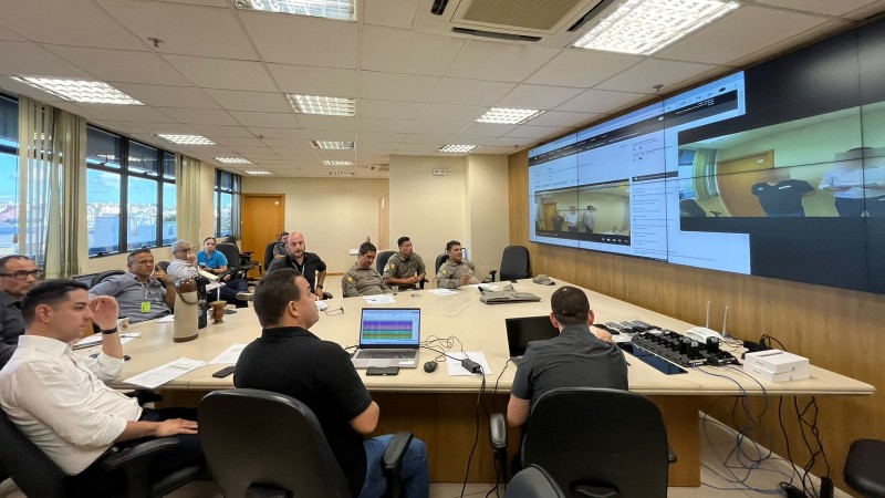 Foto em uma sala de reuniões, com uma mesa em formato triangular, direcionada para enormes telas no canto direito da imagem. As telas mostram imagens de câmera, com três pessoas paradas, com efeito blur em seus rostos. Presentes na sala, sentados em torno da mesa triangulas, há diversas autoridades, olhando para a tela à frente. Em cima da mesa, há notebooks e documentos diversos.