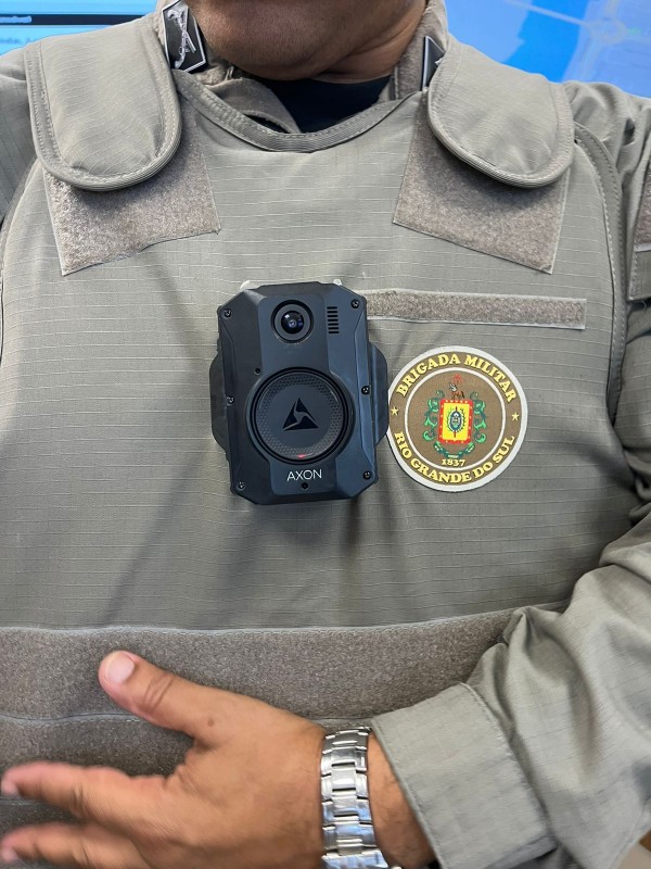 Imagem foca na câmera corporal sendo usada por um policial militar. Ela está instalada ao centro do peito, por cima do colete, que possui a marca da Brigada Militar do Rio Grande do Sul ao lado direito da foto. Não aparece o rosto do policial militar.